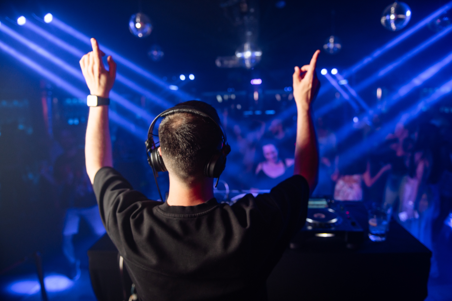 DJ with hands raised behind the decks under blue laser beams and disco balls at a NEBO night